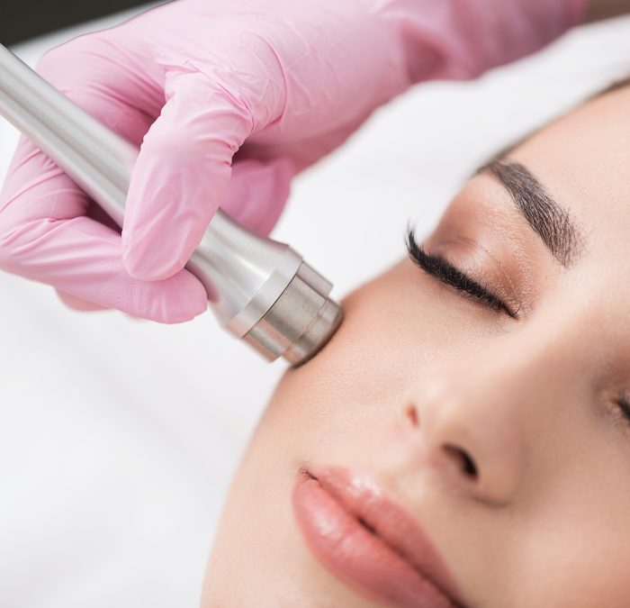 Close up of instrument in hand of doctor-cosmetologist, which is making procedure microdermabrasion of facial skin for beautiful young woman at beauty salon. Girl is lying with closed eyes